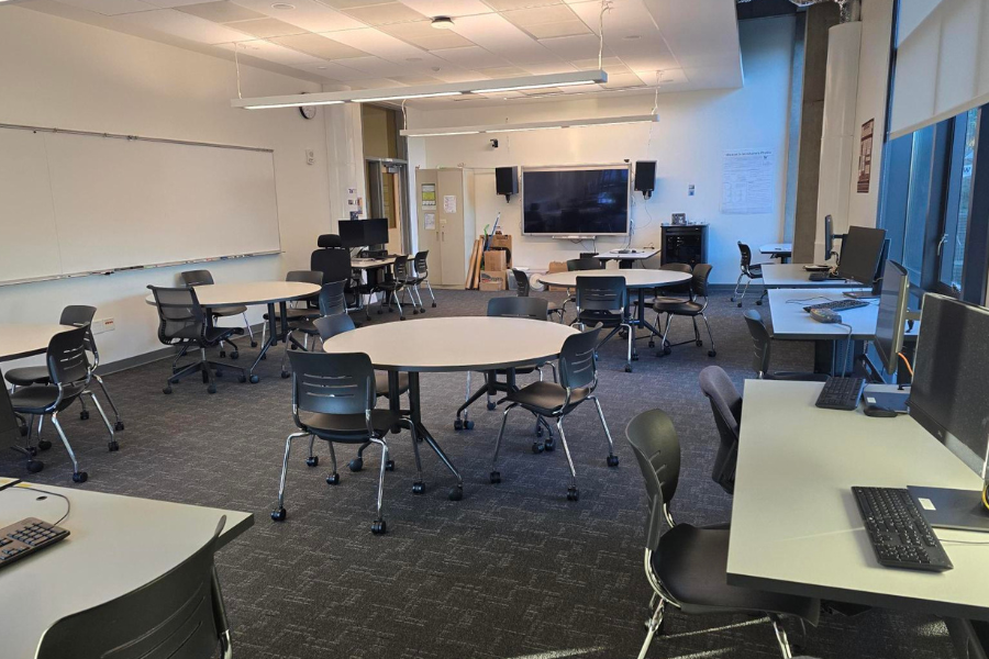 The Computational Research Lab with four round tables and rolling chairs, a large front monitor with speakers, a whiteboard along one wall, and desktop computers arranged around the remaining walls.