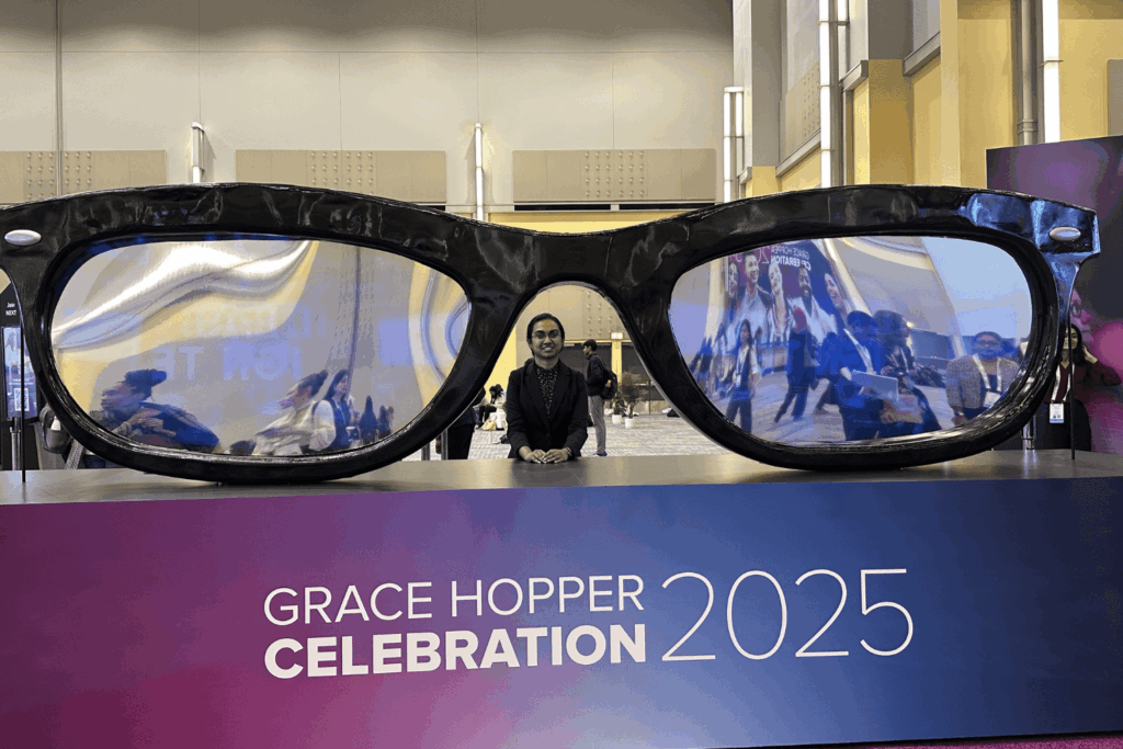 Chandramathi Murugadass at GHC 25, standing in front of a display of giant eyeglasses, with a “GRACE HOPPER CELEBRATION 2025” sign.