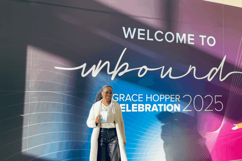 Sarah Ramazani at GHC 25, holding up her name badge in front of a sign that reads “WELCOME TO UNBOUND GRACE HOPPER CELEBRATION 2025."