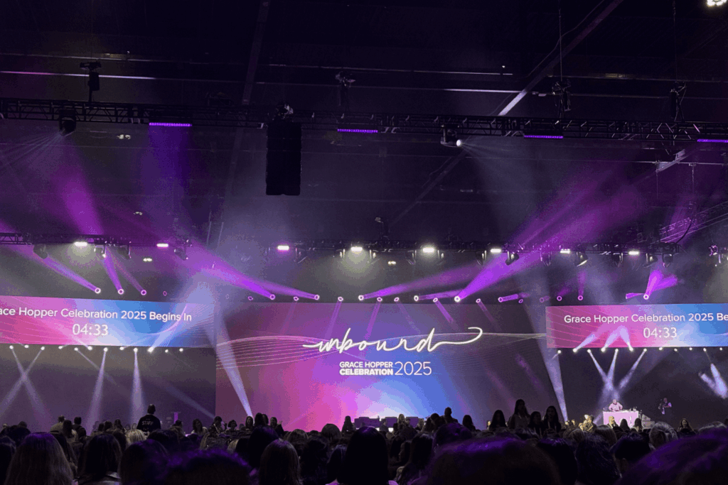 Welcome stage at GHC 25. Center screen reads “UNBOUND GRACE HOPPER CELEBRATION 2025.” Left and right screens read “Grace Hopper Celebration 2025 Begins in 04:33.” A large crowd gathers in front of the screens.