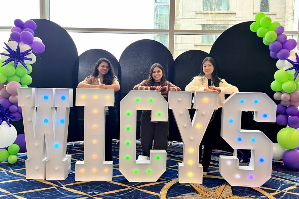 Three students standing behind a marquee display of large letters spelling ‘WICYS.’ On the sides of the display are purple/white/green balloon arches.