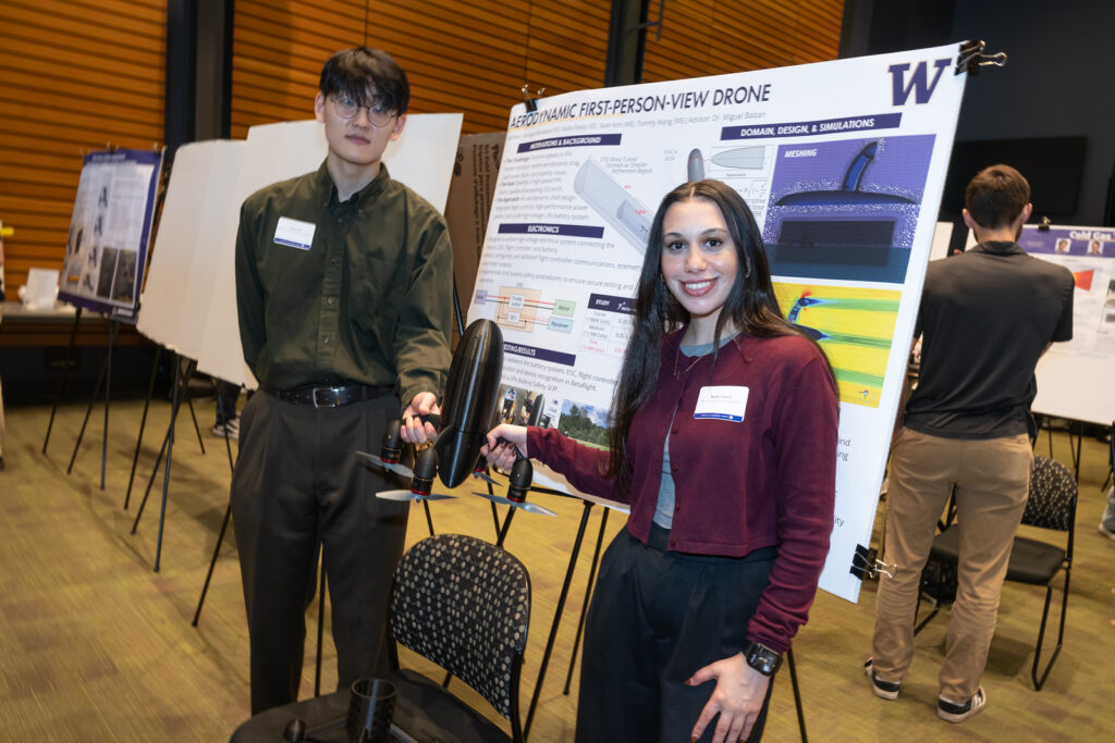 Two students stand in front of their poster board at the symposium, holding their model drone