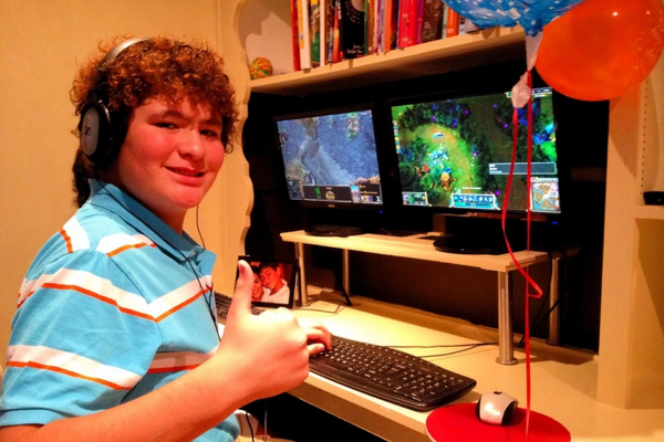 Young Bruno smiles and gives a thumbs up as he sits at a desk, playing a computer game.