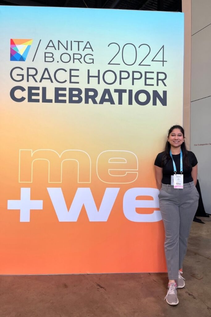 Jui stands next to an orange sign that reads "ANITA B. ORG 2024 GRACE HOPPER CELEBRATION - me + we"