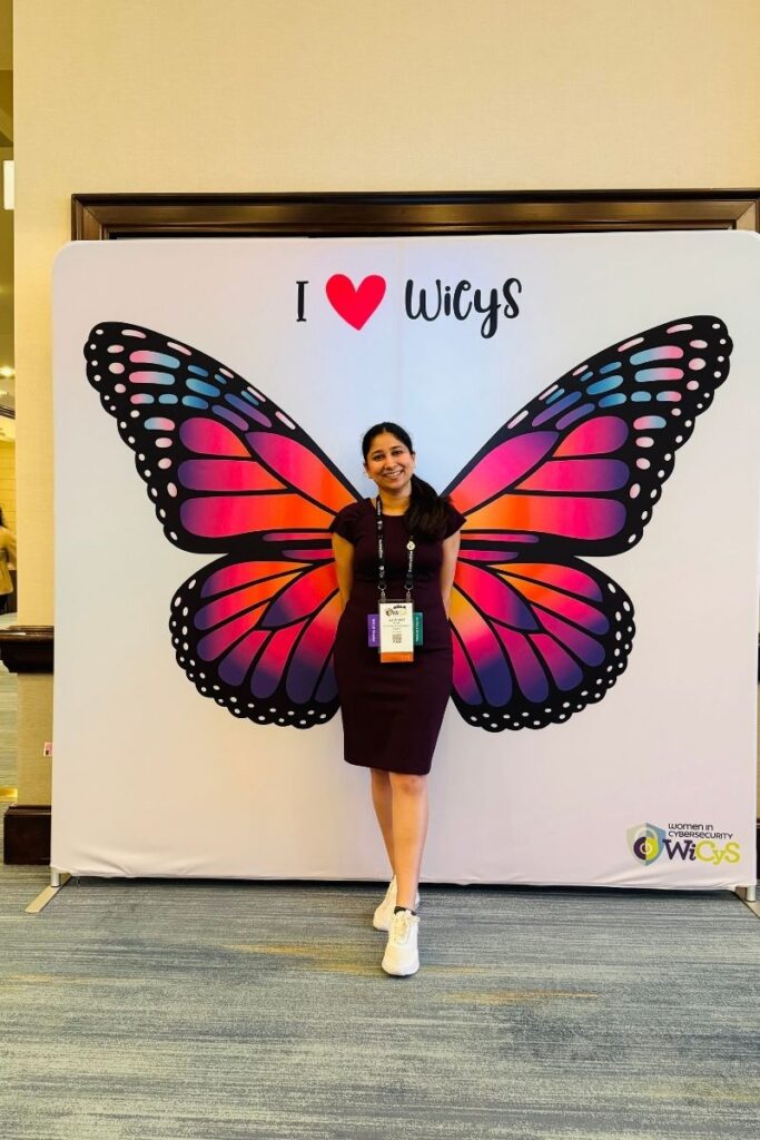 Jui stands in front of a banner that reads "I ❤︎ WiCyS” with a large pink butterfly image below