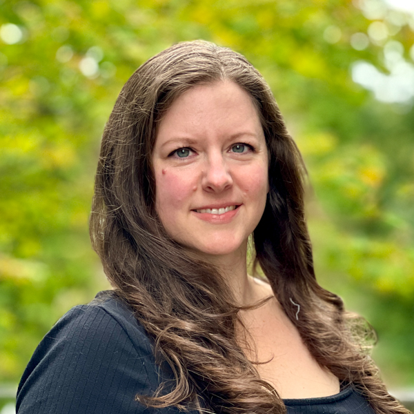 White woman with long wavy brown hair and blue eyes who is wearing a black dress.