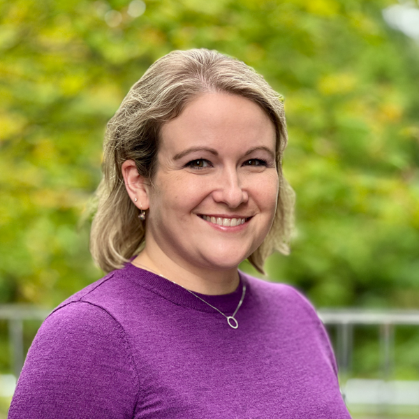 White woman with brown eyes and short blond hair wearing a purple sweater.