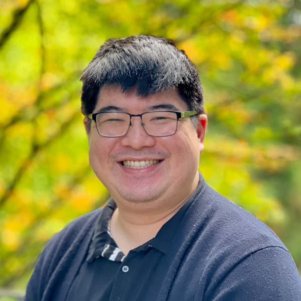 Japanese man with short black hair and brown eyes who is wearing a black collared shirt and glasses. 