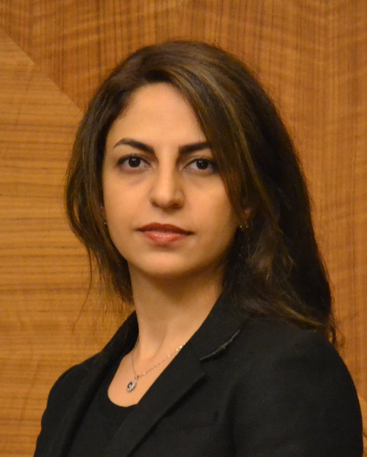 Shima Abadi headshot image with a black blazer and shirt on in front of a warm wood colored wall. She has dark brown hair and a pendant necklace on