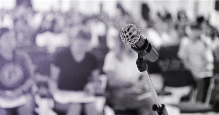 Black and white image of a microphone in focus with a blurred audience in the background 