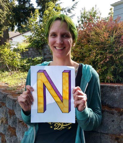 Nicole Hoover smiling and standing outside a gray brick wall in front of bushes holding a colorful yellow and purple letter “N